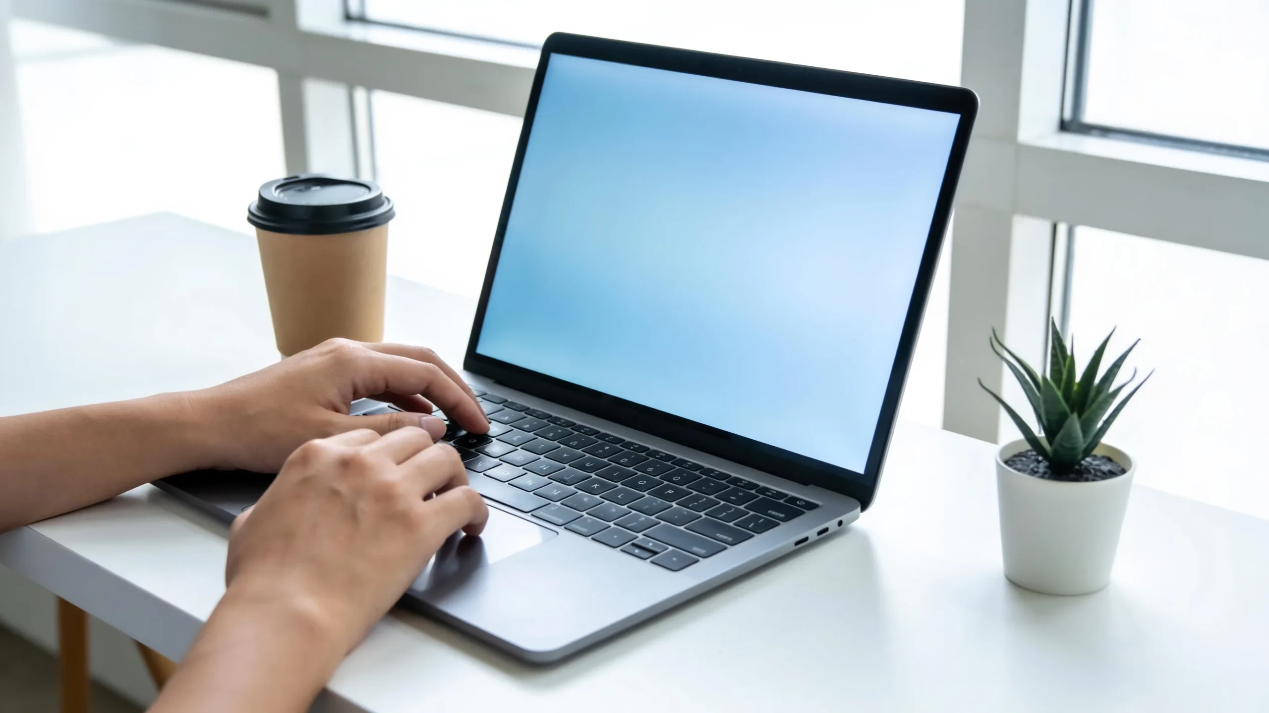Manos escribiendo un prompt en un portatil sobre un escritorio blanco con cafe y una planta