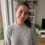 Foto de partida: retrato de medio cuerpo de una mujer con jersey gris junto a la ventana de su salón